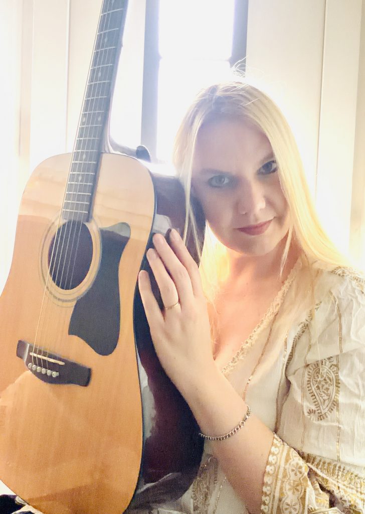 Kat Madleine is the voice of 90s revival. she is holding her acoustic guitar and looks right into the camera. The Sun is shining behind her, lighten up her blonde hair and creating an aura of pureness.
